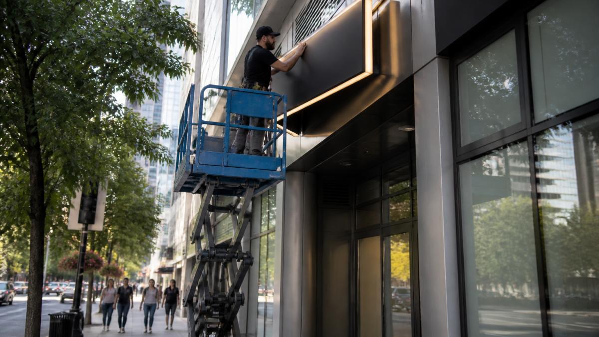A professional installer on a lift positioning a large illuminated exterior business sign on a building facade, with a street and pedestrians visible below