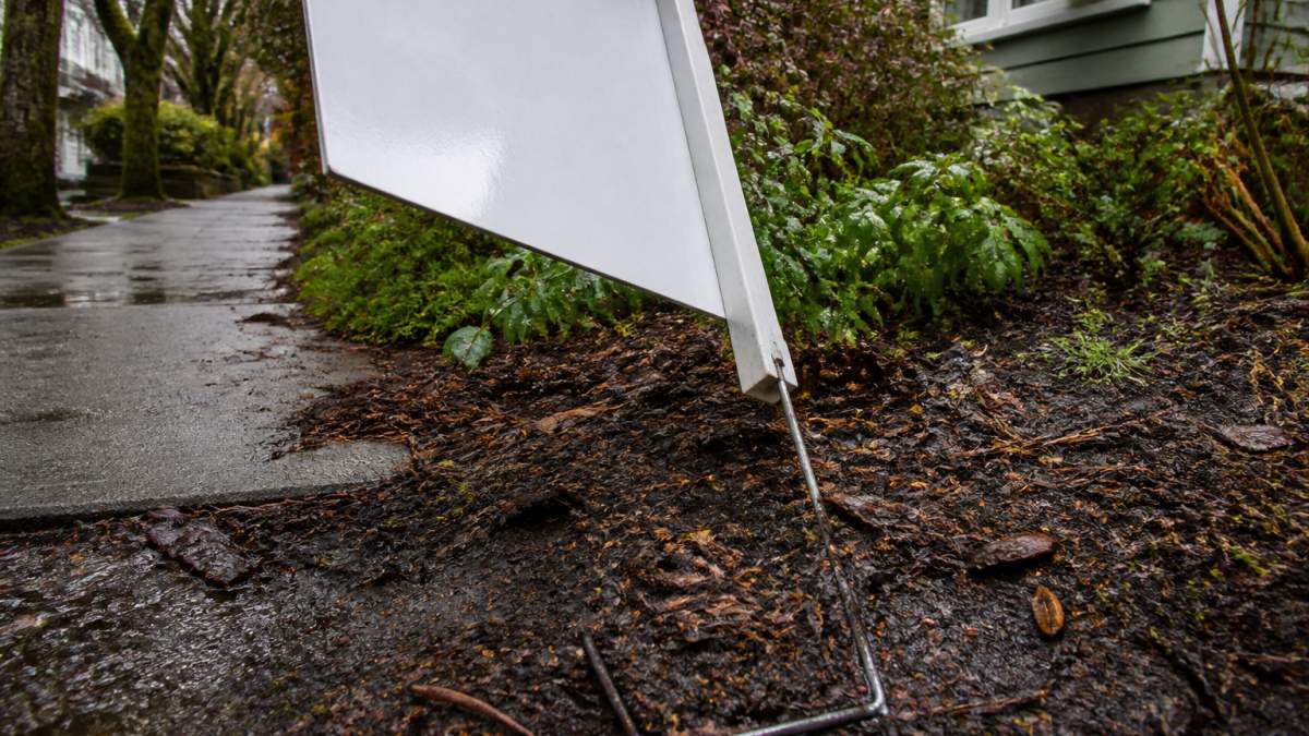 A bent metal real estate sign stake leaning in wet, muddy soil after a Pacific Northwest rainstorm.