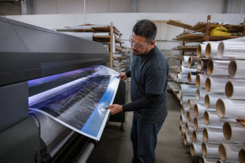 A sign shop production floor with a large-format printer running, a worker in safety glasses inspecting a vinyl print, and stacks of substrate material nearby