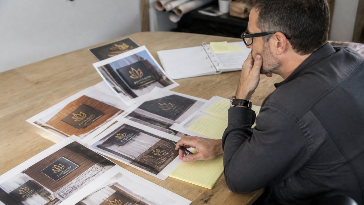 Project manager reviewing multiple versions of a digital signage design on a computer monitor, with printed design proofs and revision notes on the desk nearby