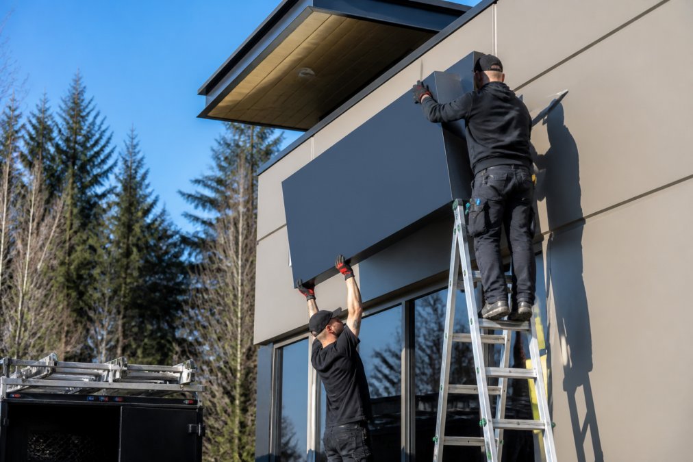 A professional installation team in branded workwear mounting a large commercial sign on the exterior of a modern building in the Pacific Northwest, with a clear blue sky and evergreen trees in the background